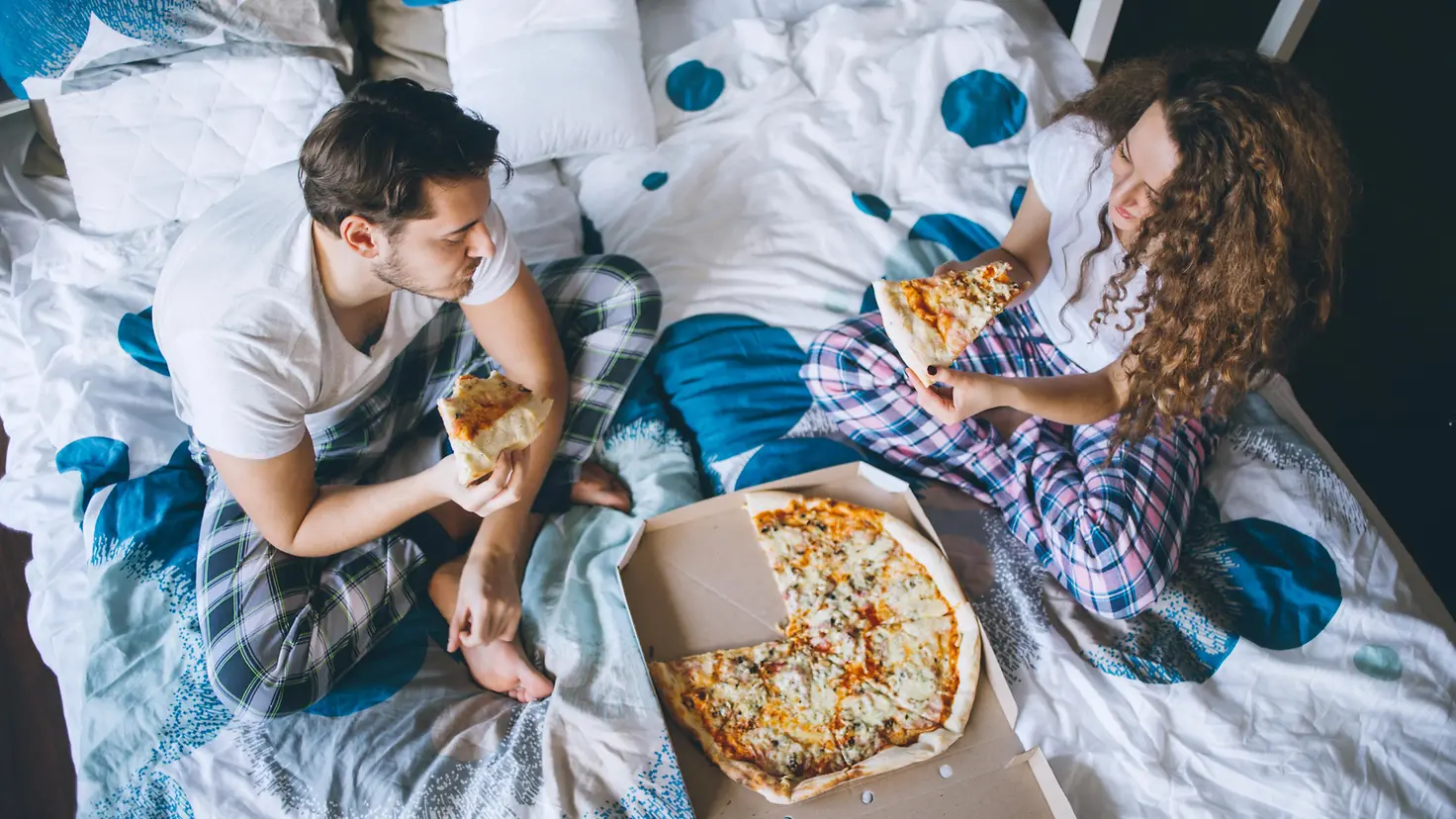 Zwei Personen sitzen auf dem Bett und essen genüsslich eine Pizza