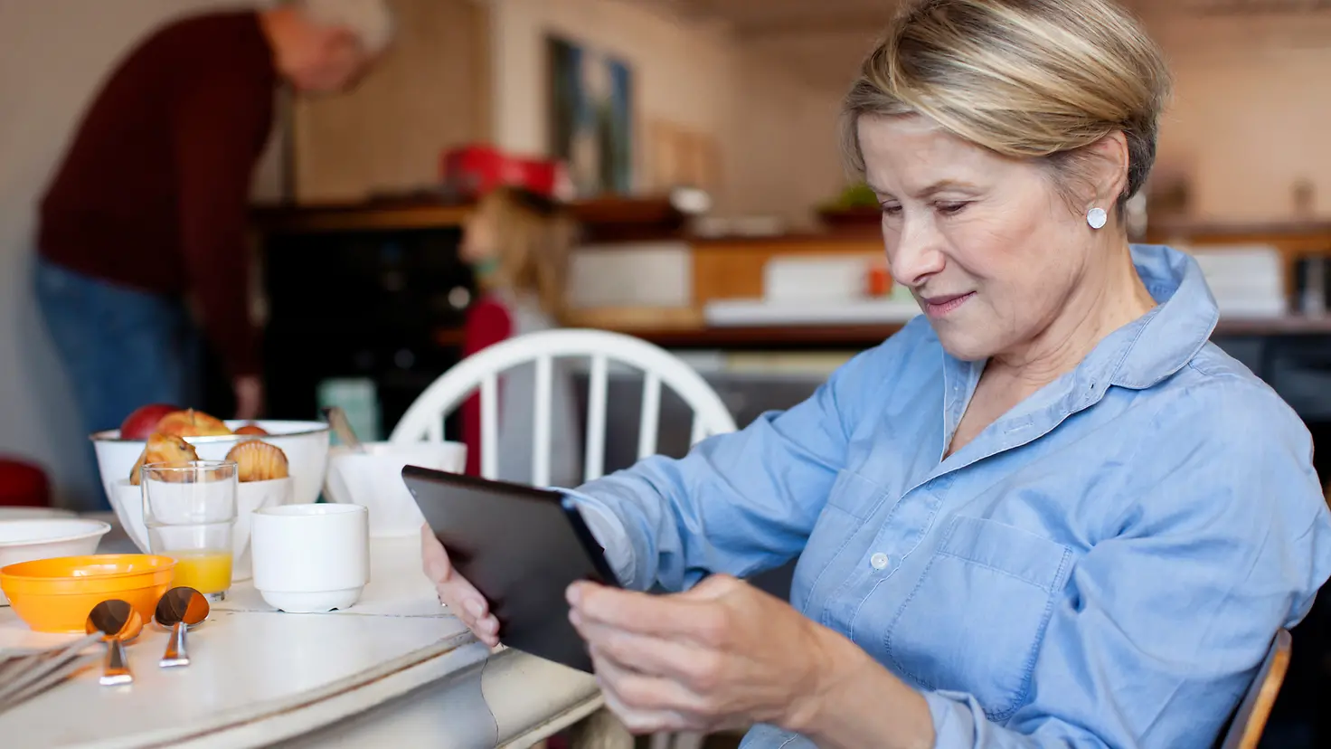 Eine ältere Frau sitzt mit Tablet am Küchentisch und informiert sich über die Krankenversicherung für Rentner. 