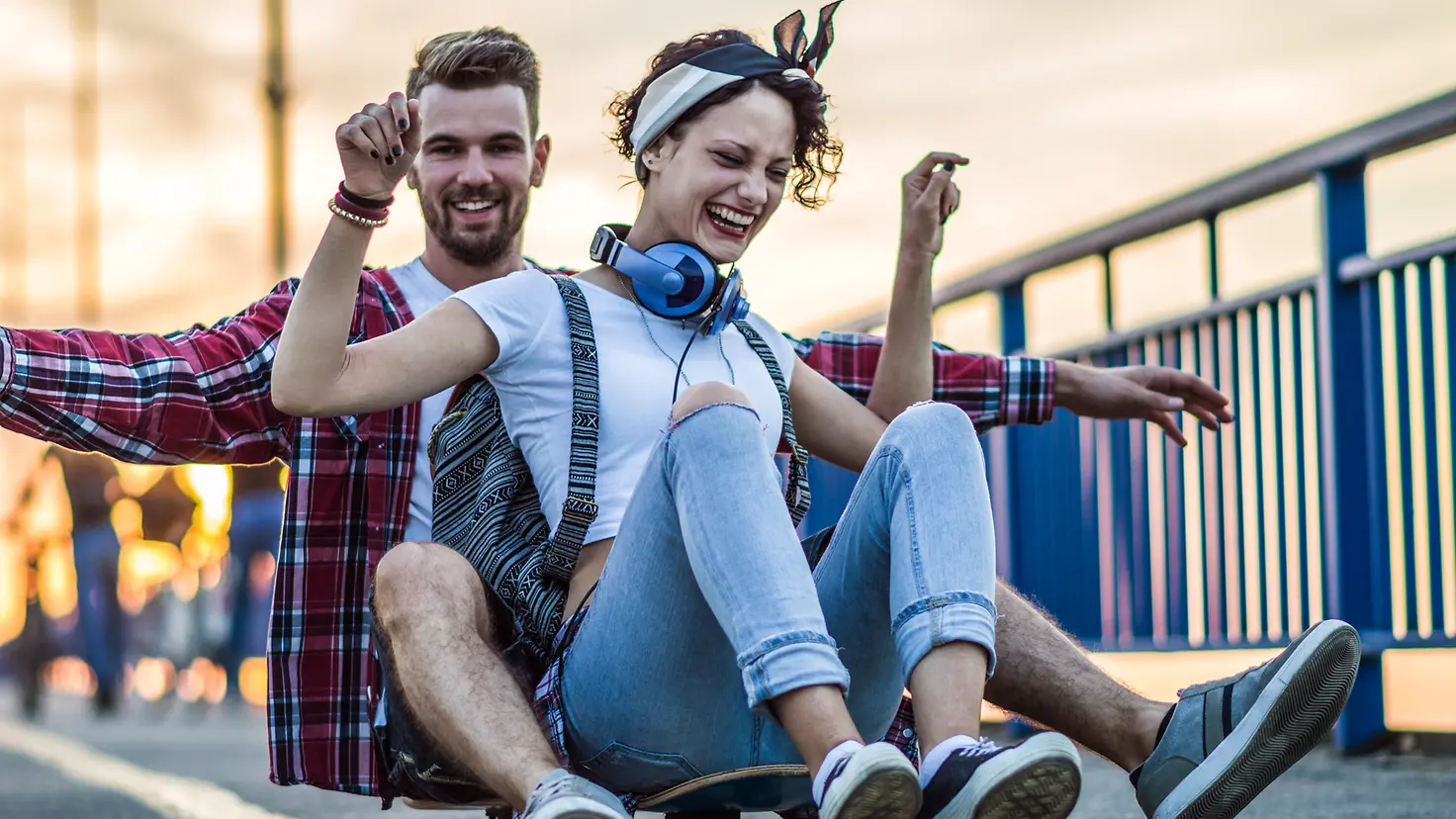 Ein junger Mann und eine extrovertierte, junge Frau sitzen lachend auf einem rollenden Skateboard.